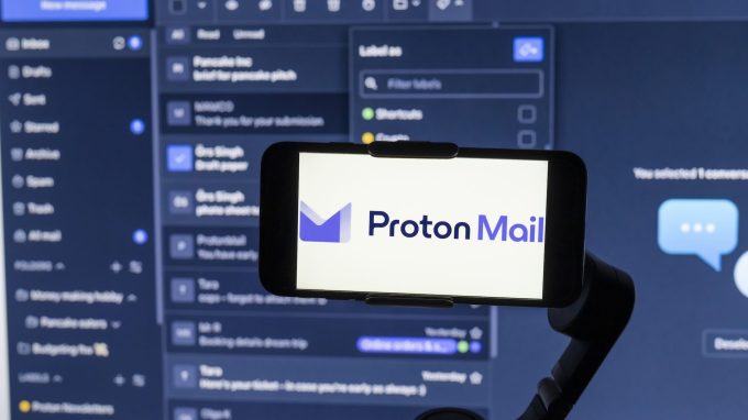 ProtonMail logo seen displayed on a mobile phone screen with its website interface in the background.