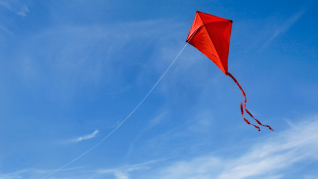 A photograph of a red kite flying under a blue sky with a few whisps of clouds.