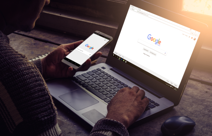 A person using a smartphone and laptop with Google homepage on display.