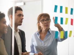 Boring governance is the trail to actual AI adoption Middle aged businesswoman mentor coach leader explaining project strategy showing post it sticky notes on glass wall, serious business team brainstorm work plan at corporate office stand up meeting