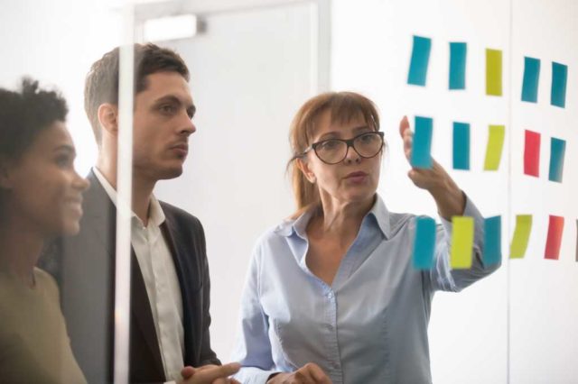 Boring-governance-is-the-path-to-real-AI-adoption.jpg Middle aged businesswoman mentor coach leader explaining project strategy showing post it sticky notes on glass wall, serious business team brainstorm work plan at corporate office stand up meeting