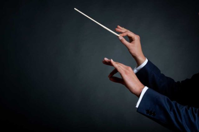 The hands of a male orchestra conductor directing with his baton in concert. Ivory baton white cuffs and dark blue suit against a dark gray background.