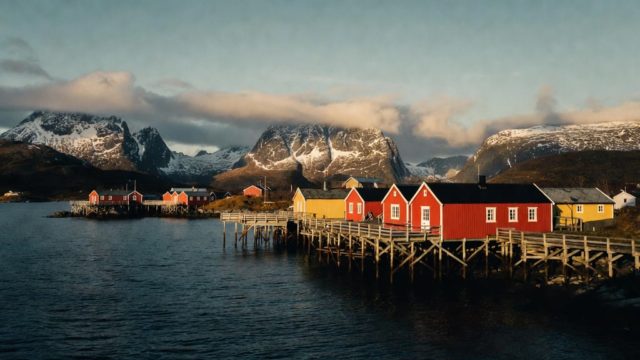 A scenery of houses along the shoreline with snowy mountains in the background.